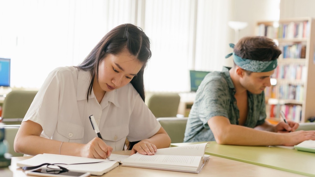 Two students writing at a shared table in a bright university library