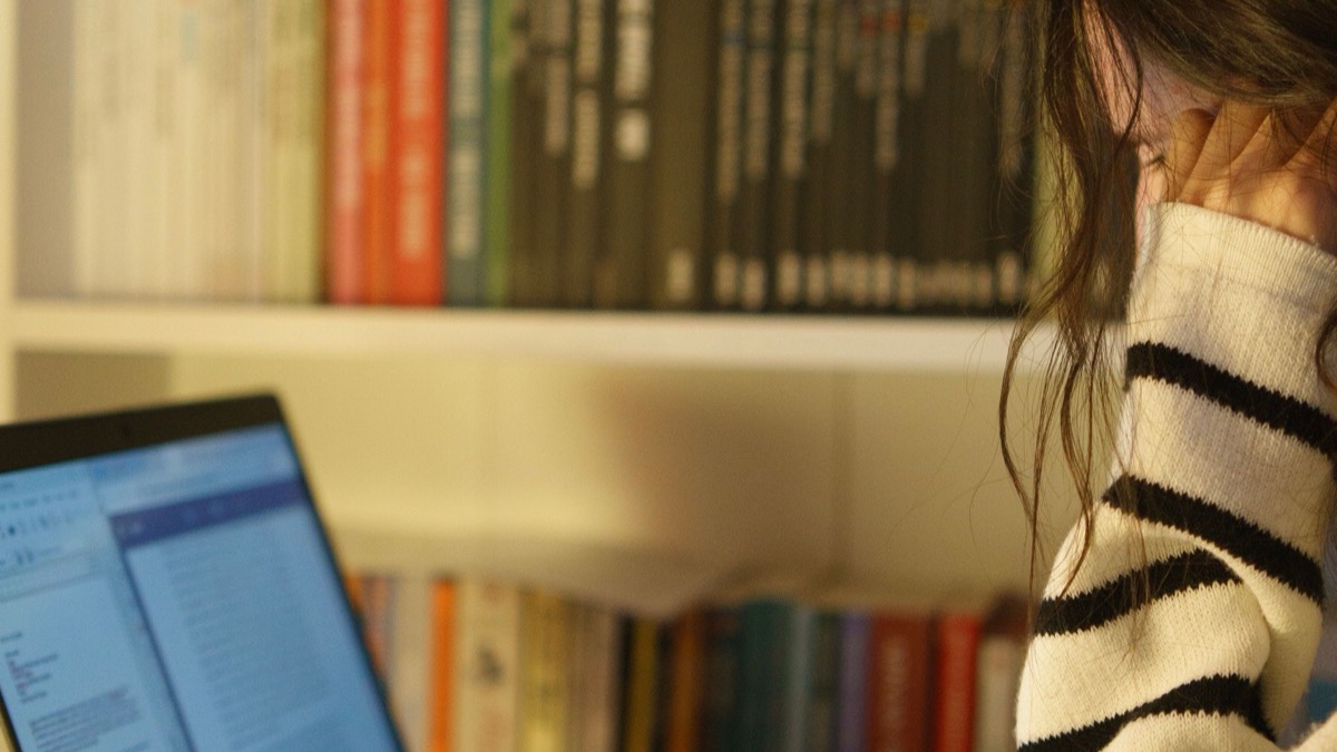A student writing at a laptop beside a bookshelf